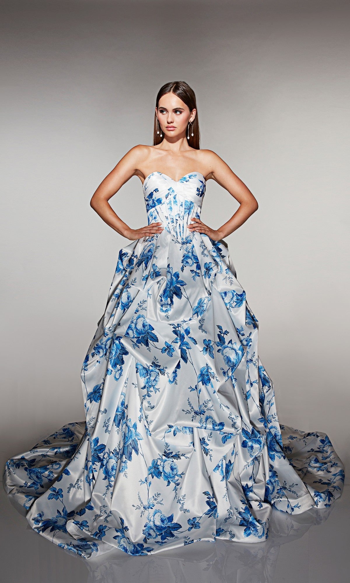 A woman stands with hands on hips, wearing the Long Prom Dress: Alyce 62213 in Blue/White, featuring a corset bodice, strapless neckline, and a long flowing train against a plain background.